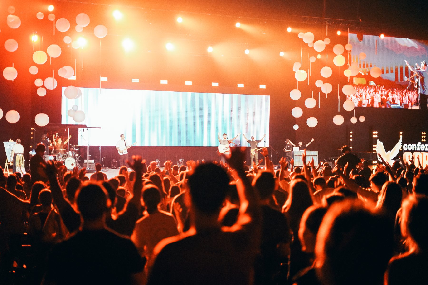 Plateia de braços erguidos durante um show, com o palco iluminado de laranja ao fundo