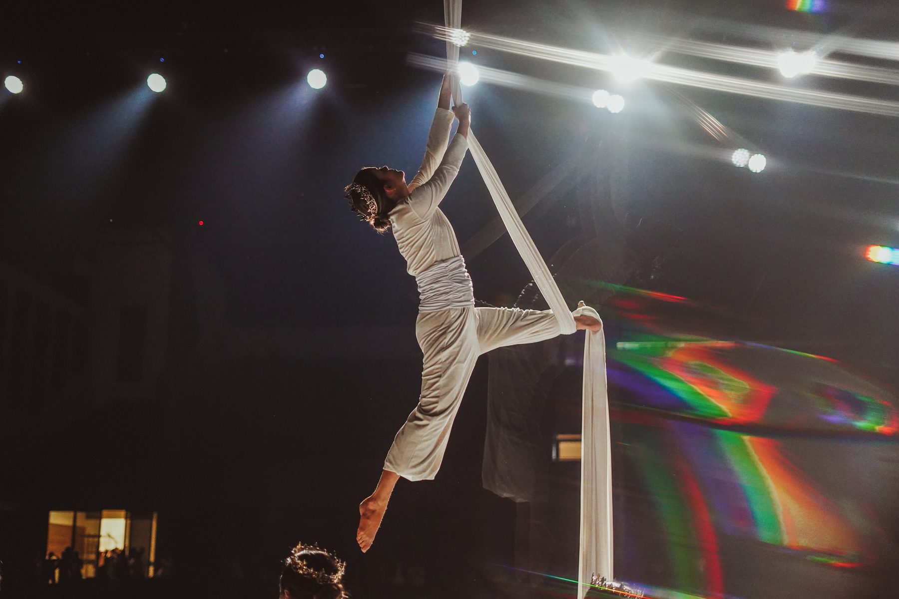 Performer suspensa em tecidos aéreos no palco, com luzes coloridas ao fundo