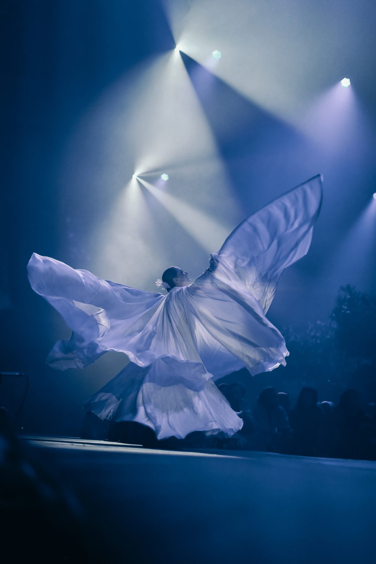Dançarina com grandes asas brancas em pleno movimento, luz azul de palco