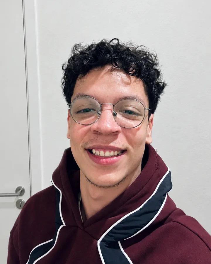 Retrato de Leonardo Martinelli sorrindo, com cabelo cacheado, óculos redondos e moletom vinho.